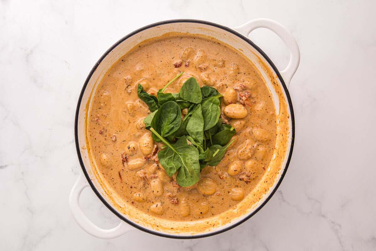 Fresh baby spinach added on top of gnocchi and creamy sauce in a white skillet before wilting.