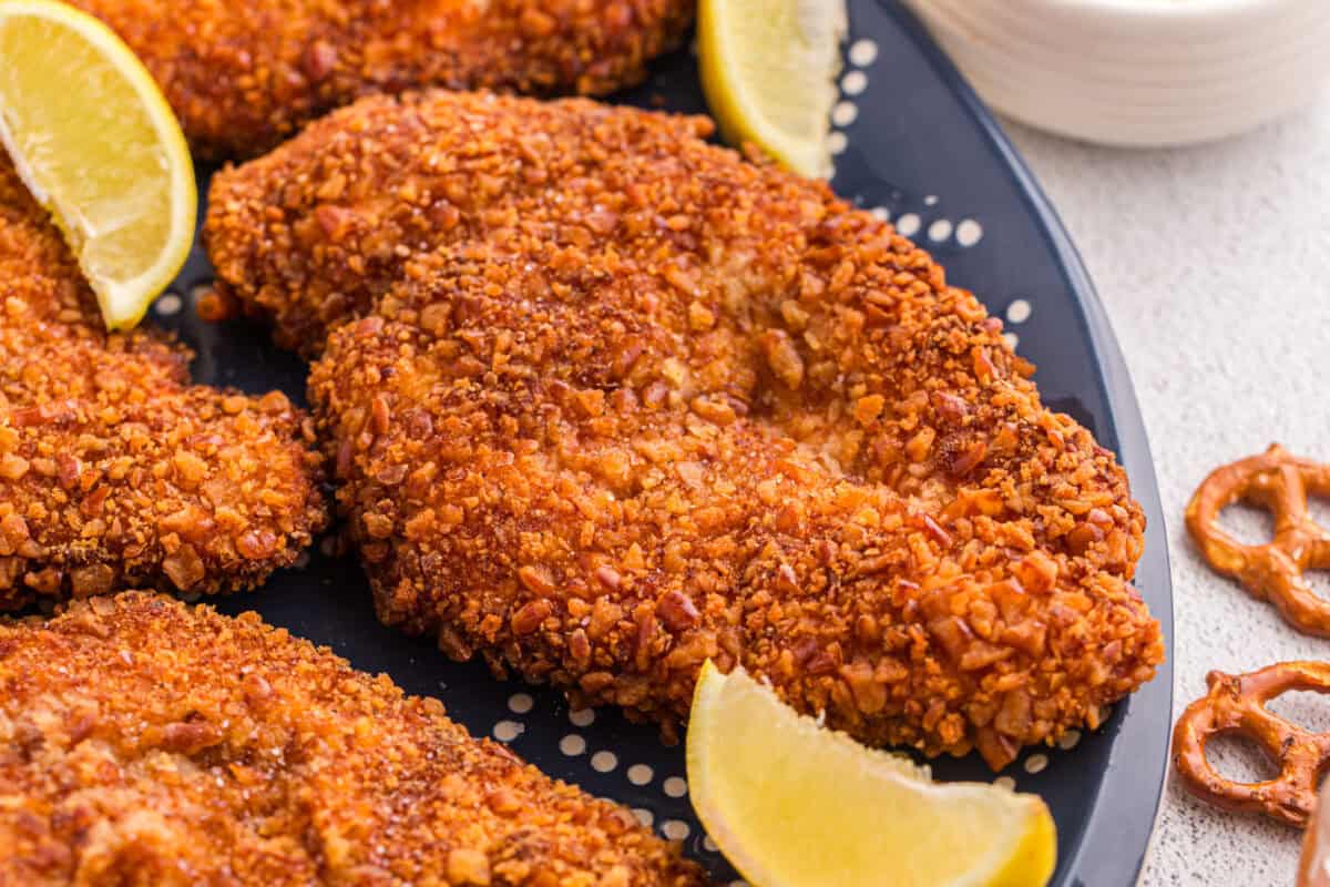 Close up of pretzel crusted chicken cutlets on a dark blue platter showing the crunchy golden brown coating with lemon wedges
