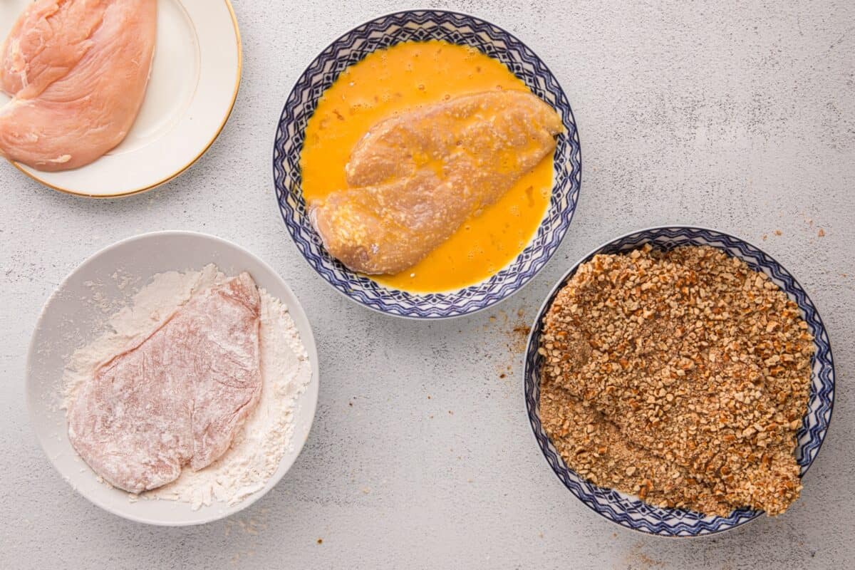 Chicken cutlet being dredged through flour, egg wash, and pretzel coating bowls in the three step breading process