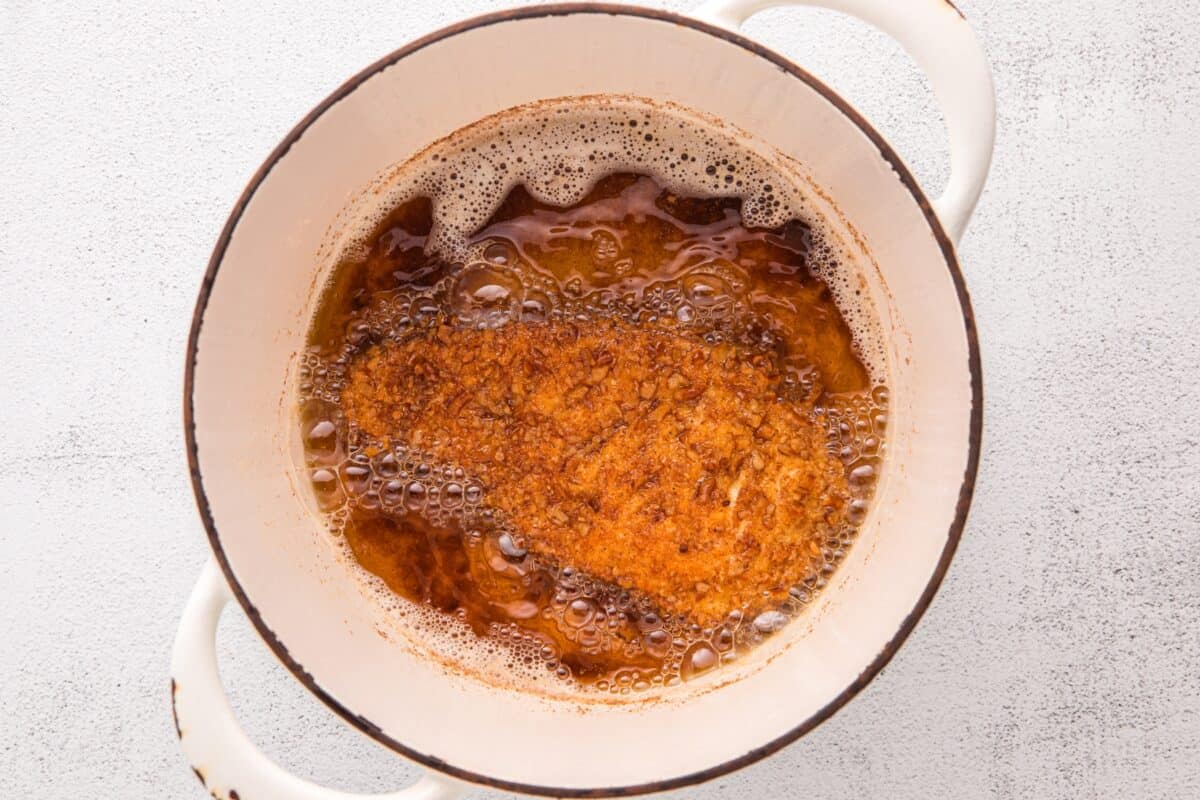 Pretzel crusted chicken cutlet frying in hot oil in a white dutch oven with a golden brown crust forming