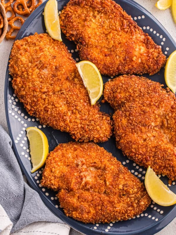 Pretzel crusted chicken cutlets on a dark blue oval platter with lemon wedges and whole pretzels in the background