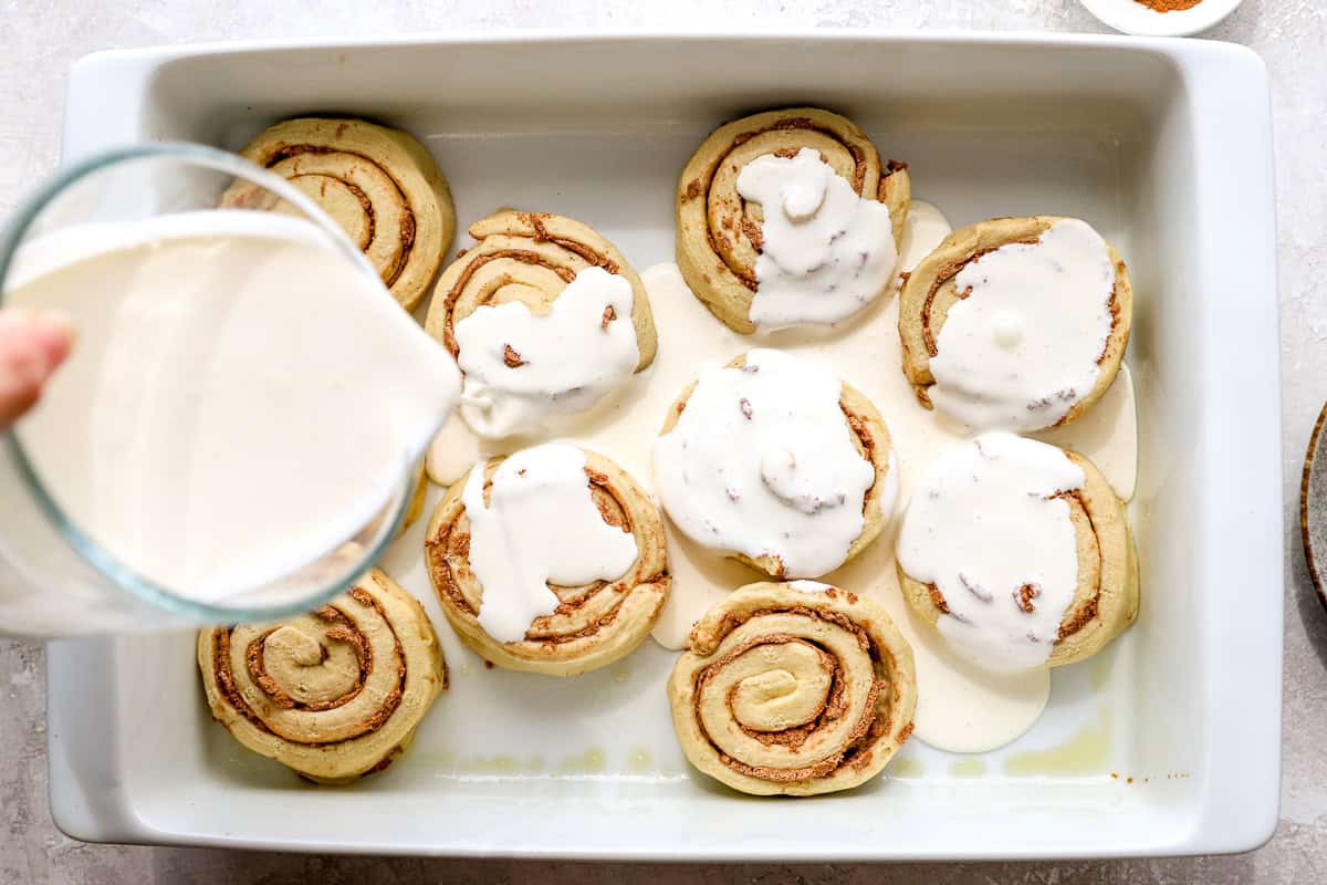 Heavy cream being poured over refrigerated cinnamon rolls in a baking dish.