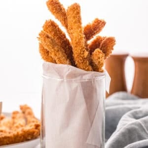 Crispy breaded chicken fries standing upright in a parchment-lined glass, with more fries and dipping sauce blurred in the background.
