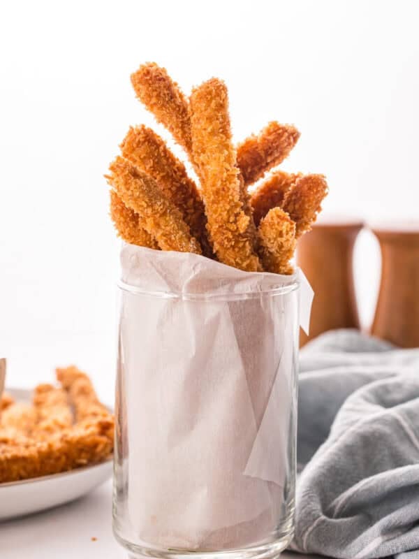 Crispy breaded chicken fries standing upright in a parchment-lined glass, with more fries and dipping sauce blurred in the background.