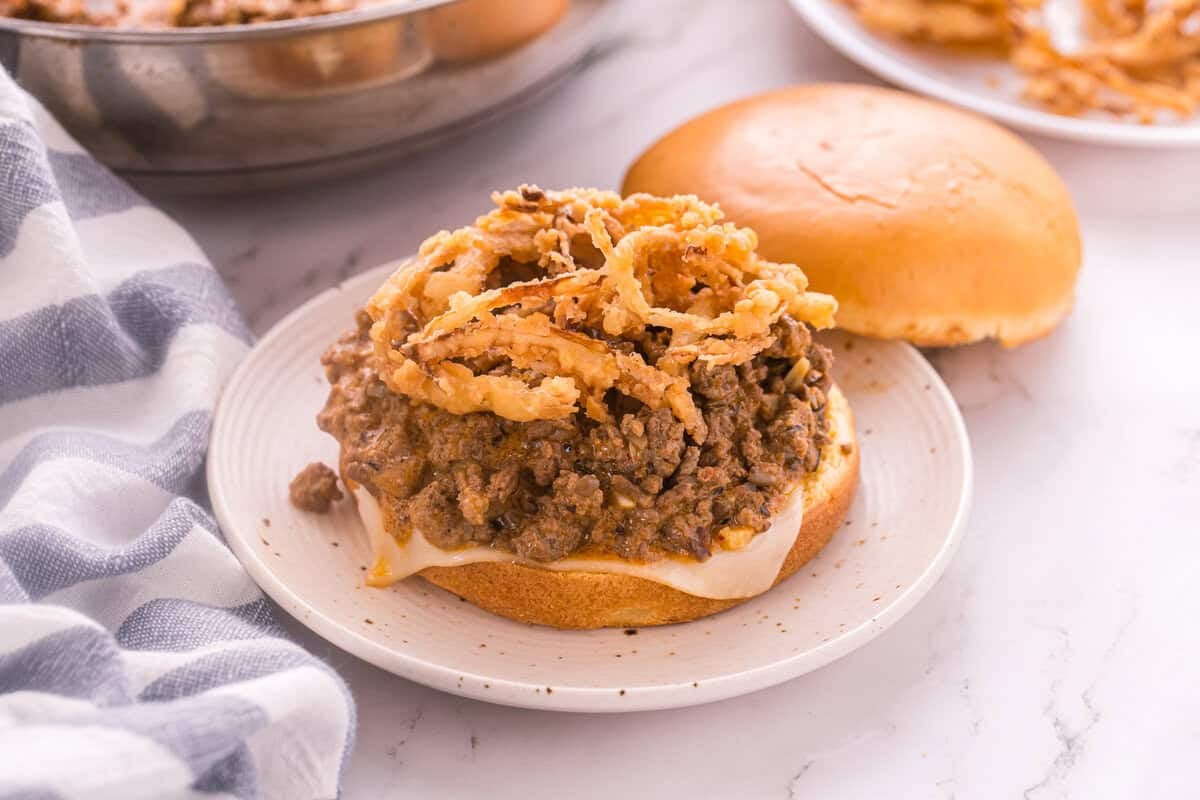 Bottom brioche bun topped with melted Swiss cheese, beef stroganoff filling, and crispy fried onions next to the top bun