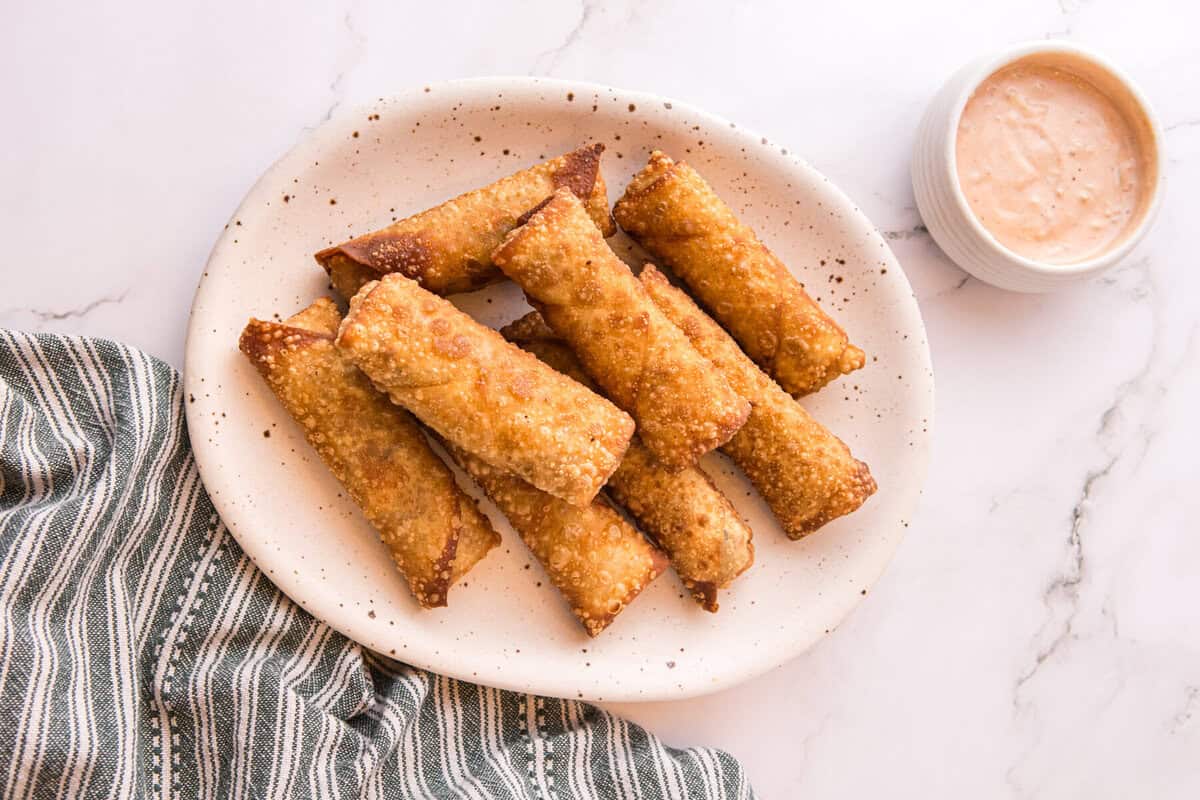 Golden fried Reuben egg rolls arranged on an oval plate next to a bowl of creamy dipping sauce.