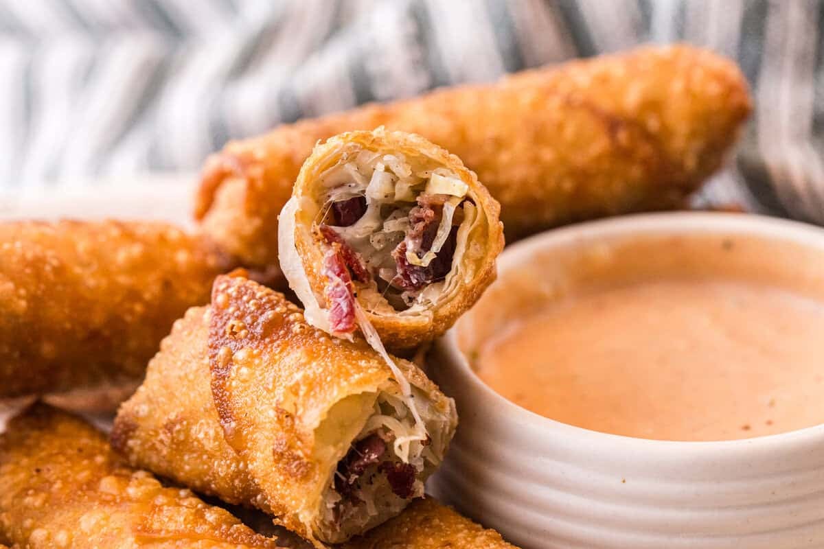 A Reuben egg roll sliced open to show the melty Swiss cheese, corned beef, and sauerkraut filling, served with dipping sauce.