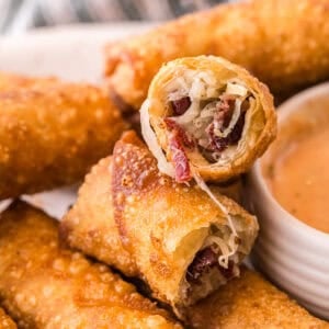 A close-up of a Reuben egg roll cut open, showing melty Swiss cheese, corned beef, and sauerkraut inside the crispy fried shell, with a bowl of dipping sauce beside it.