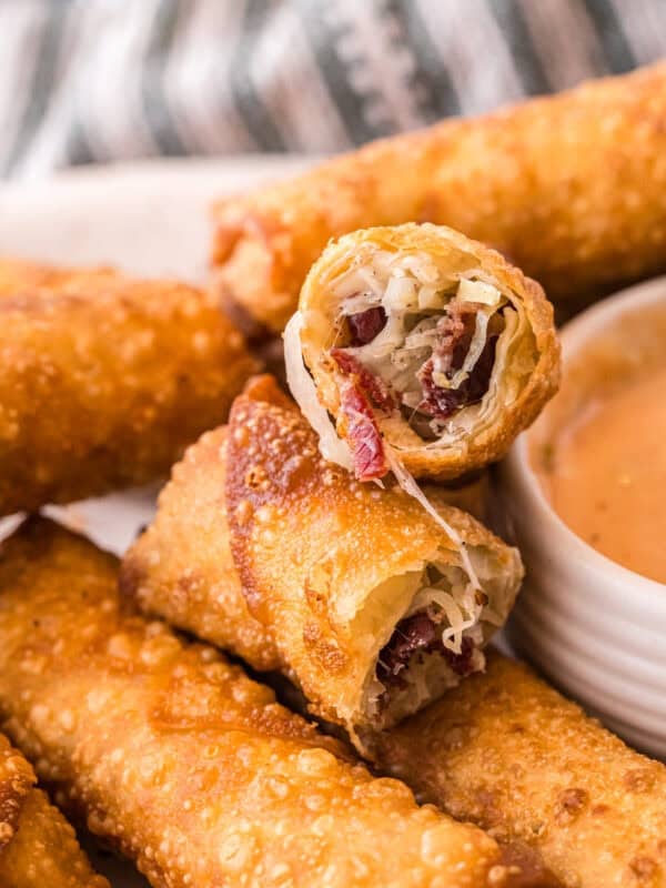 A close-up of a Reuben egg roll cut open, showing melty Swiss cheese, corned beef, and sauerkraut inside the crispy fried shell, with a bowl of dipping sauce beside it.
