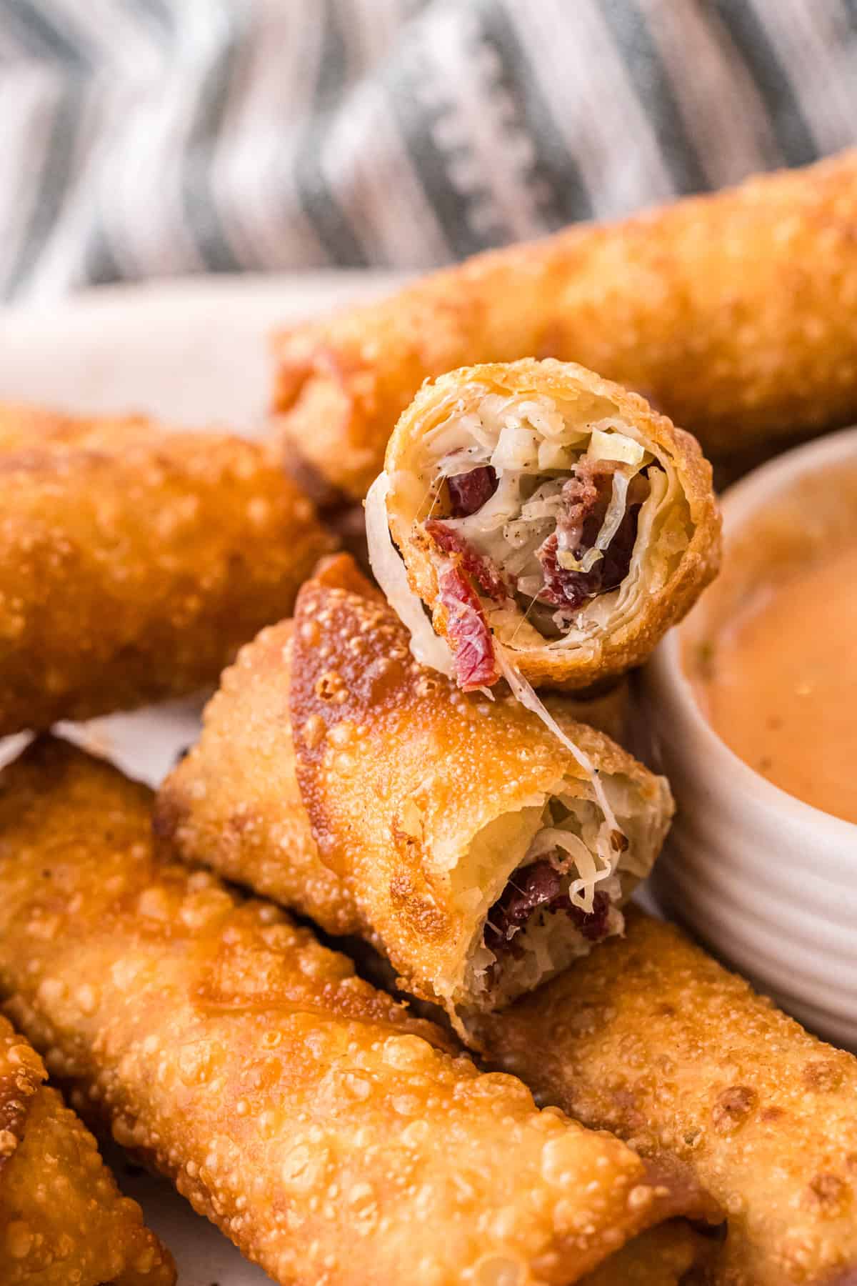A close-up of a Reuben egg roll cut open, showing melty Swiss cheese, corned beef, and sauerkraut inside the crispy fried shell, with a bowl of dipping sauce beside it.