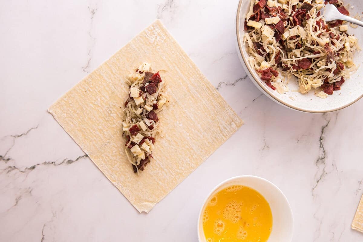 Reuben filling spooned onto the center of an egg roll wrapper, with a bowl of beaten egg for sealing nearby.