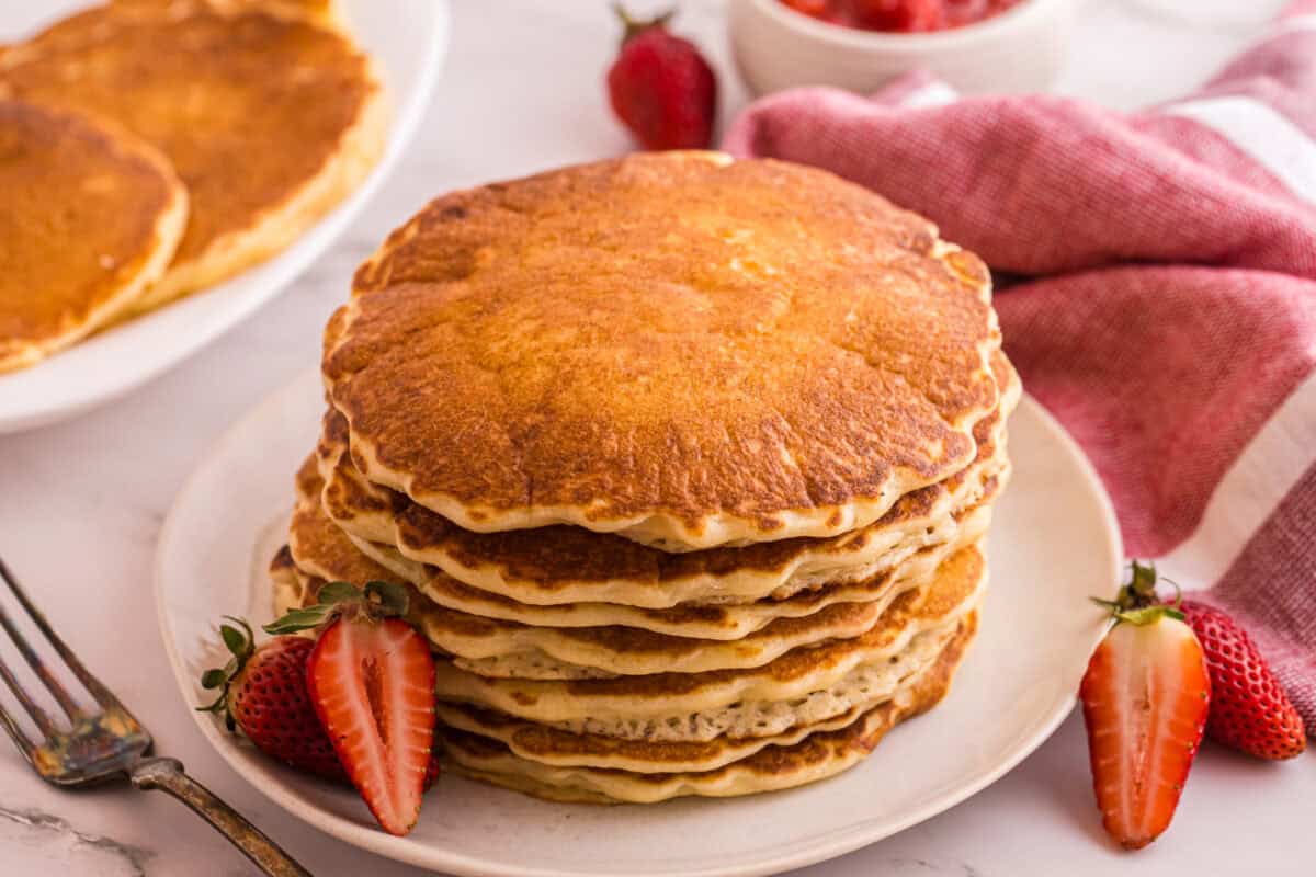 A stack of golden brown pancakes on a white plate with fresh strawberries, before topping