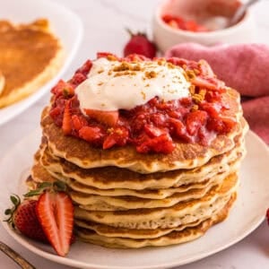 Stack of strawberry cheesecake pancakes topped with strawberry sauce, whipped cream, and graham cracker crumbs on a white plate