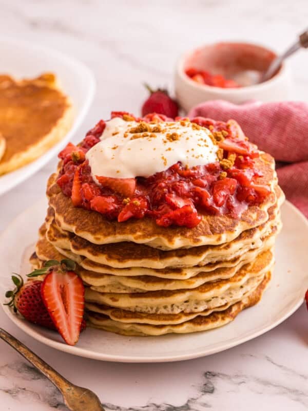 Stack of strawberry cheesecake pancakes topped with strawberry sauce, whipped cream, and graham cracker crumbs on a white plate
