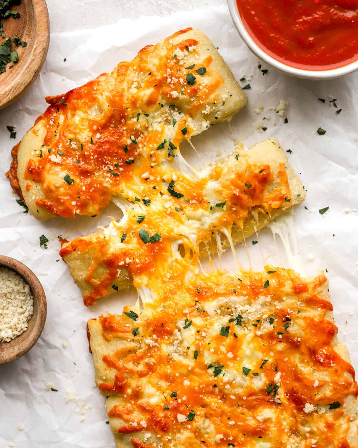 Overhead shot of sliced stuffed cheesy bread with melted cheese pulling apart, next to a bowl of marinara sauce and grated parmesan