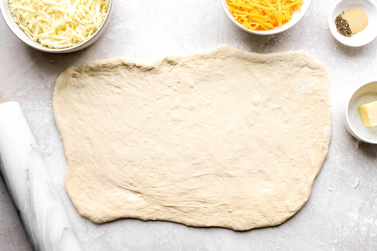 Pizza dough rolled into a large rectangle on a floured surface next to bowls of shredded mozzarella, cheddar, and garlic herb seasoning