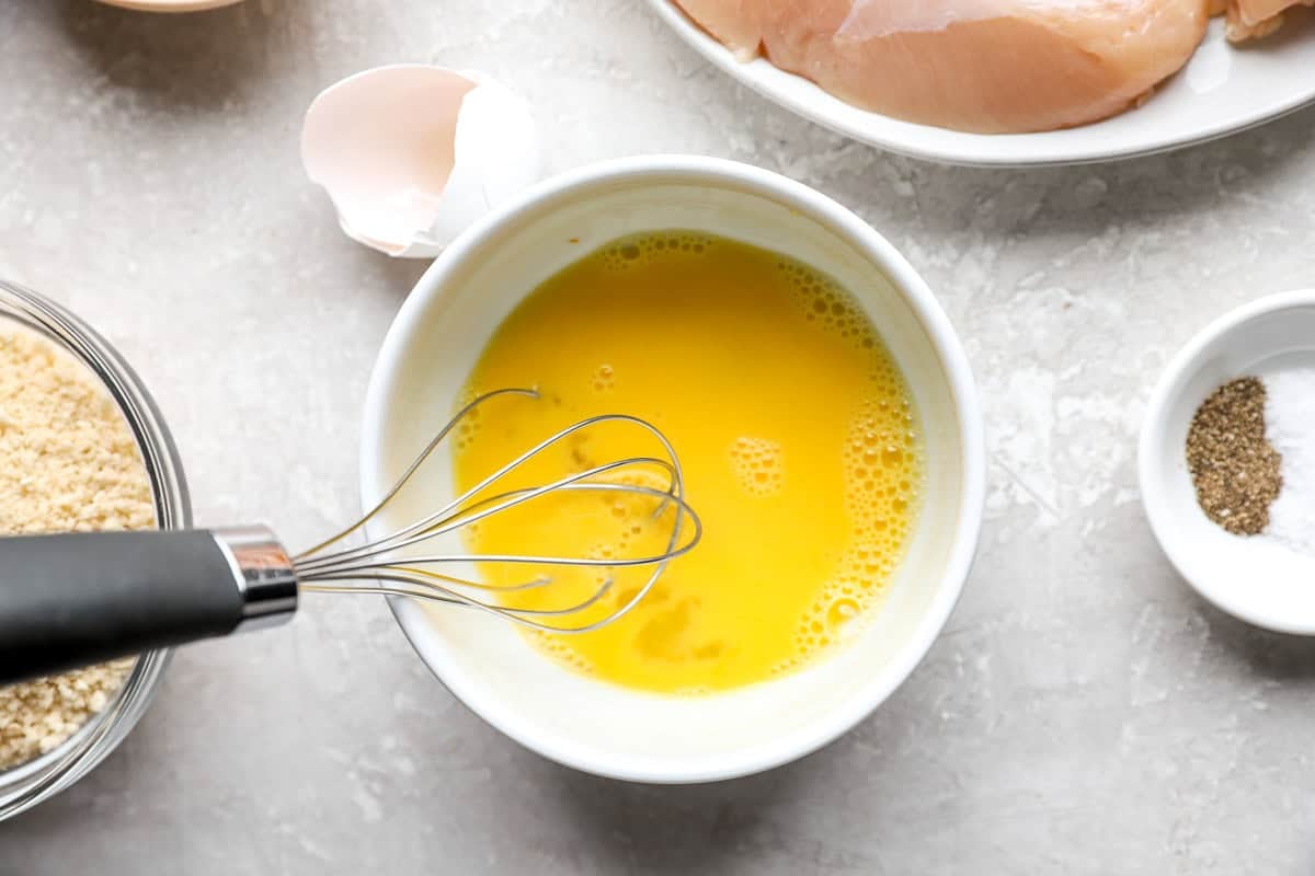 Whisked egg in a white bowl with a whisk, raw chicken tenders and a cracked eggshell in the background