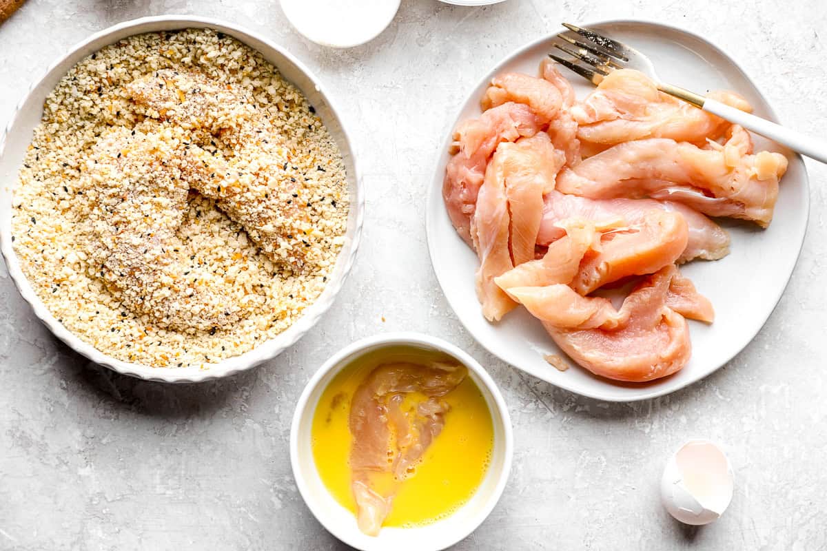 Raw chicken tender being pressed into a bowl of panko and everything bagel seasoning, with more tenders and egg wash on the side.