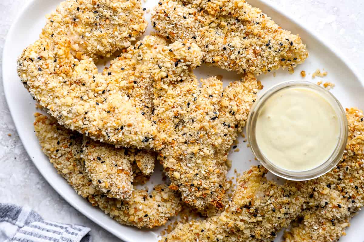 Crispy everything bagel chicken strips piled on a white platter next to a bowl of dipping sauce.