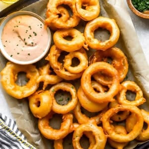 Overhead shot of crispy homemade onion rings piled on a parchment-lined tray with a bowl of pink dipping sauce, a bowl of chopped herbs, and a glass of beer alongside.