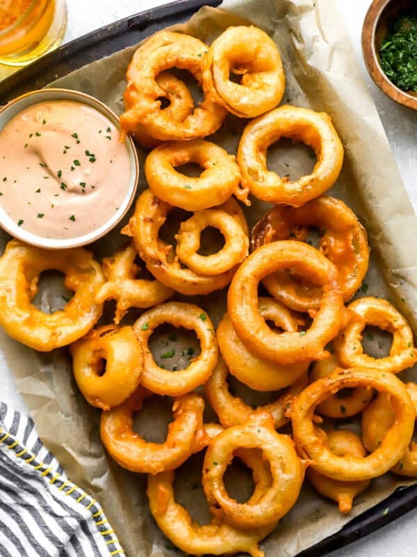 Overhead shot of crispy homemade onion rings piled on a parchment-lined tray with a bowl of pink dipping sauce, a bowl of chopped herbs, and a glass of beer alongside.