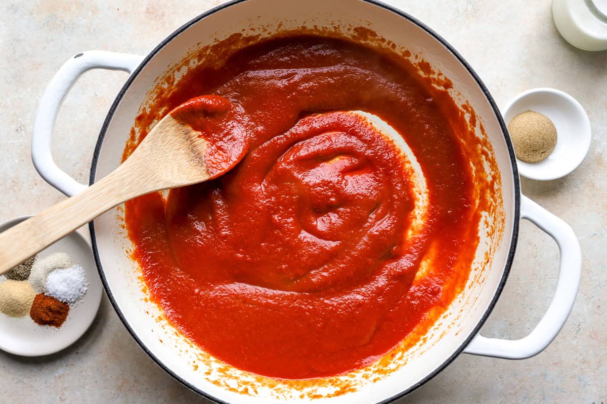 Tomato sauce and tomato paste stirred together in a white pot with a wooden spoon, before adding seasonings.