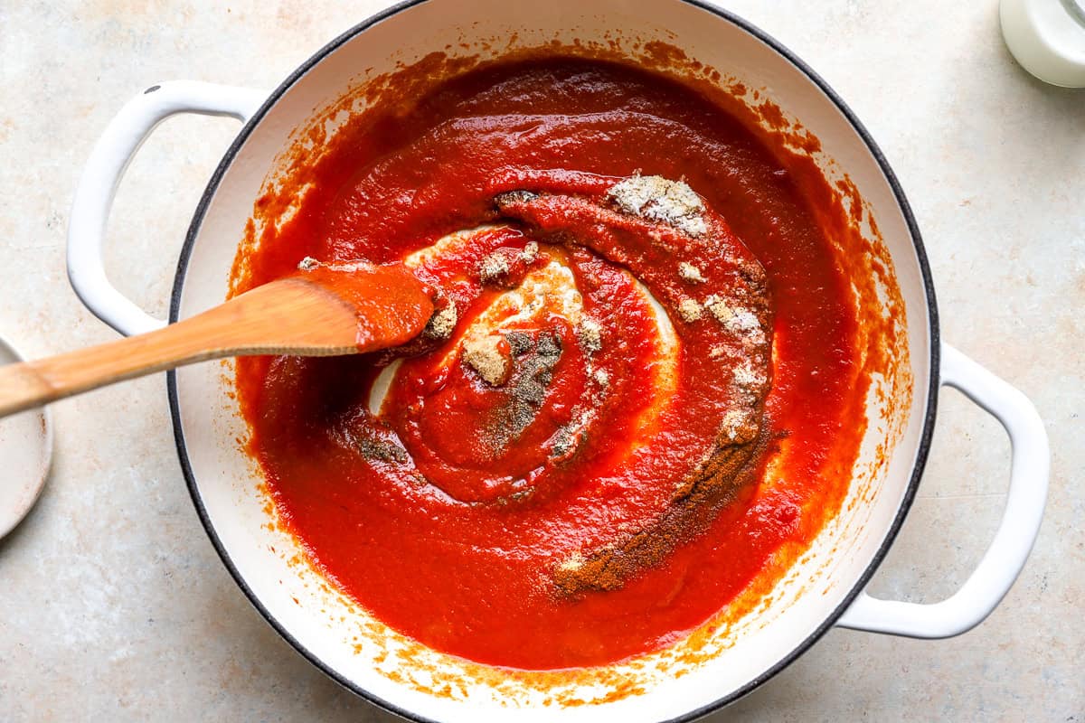 Garlic powder, onion powder, paprika, salt, and pepper being stirred into the tomato sauce in a large pot.