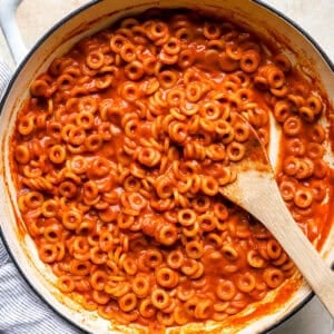 Overhead view of homemade SpaghettiOs in a white pot, coated in glossy tomato sauce with a wooden spoon resting inside.
