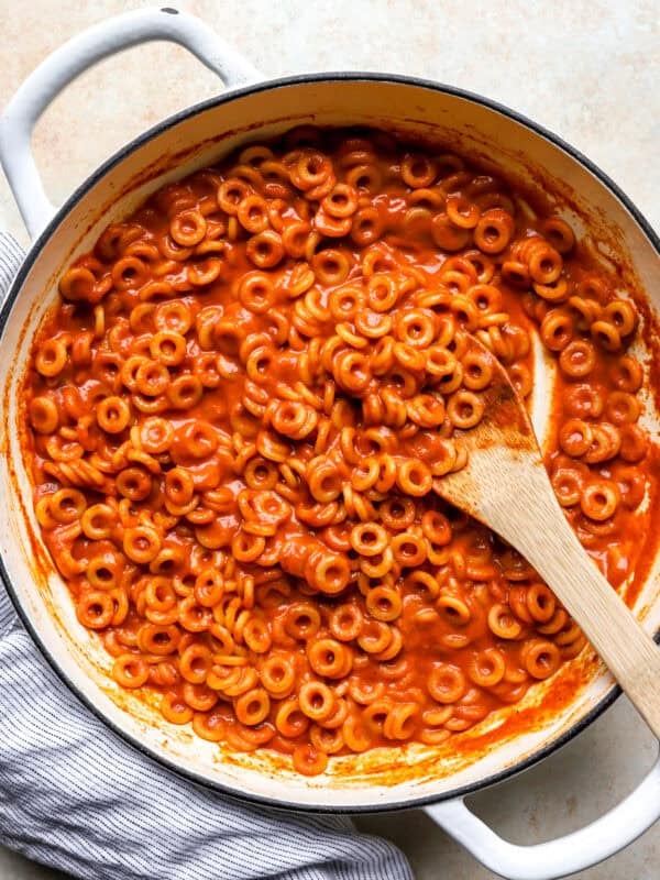 Overhead view of homemade SpaghettiOs in a white pot, coated in glossy tomato sauce with a wooden spoon resting inside.