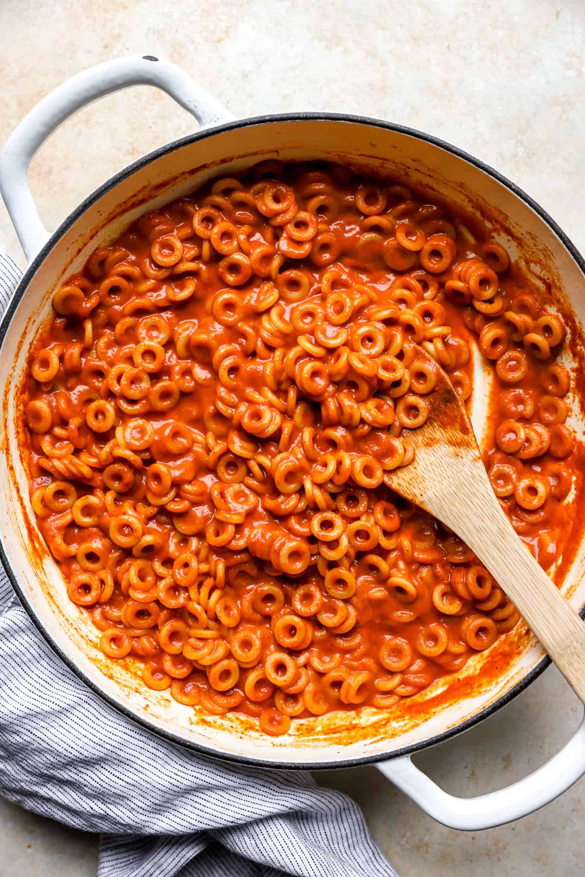 Overhead view of homemade SpaghettiOs in a white pot, coated in glossy tomato sauce with a wooden spoon resting inside.
