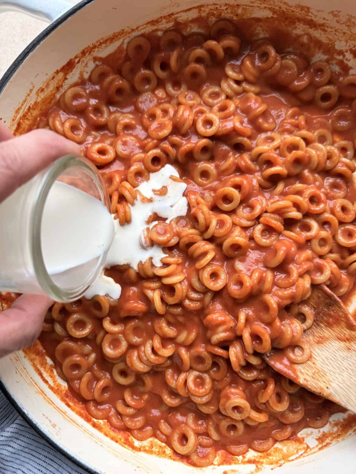 Heavy cream being poured into the pot of saucy anellini pasta to make the sauce rich and creamy.