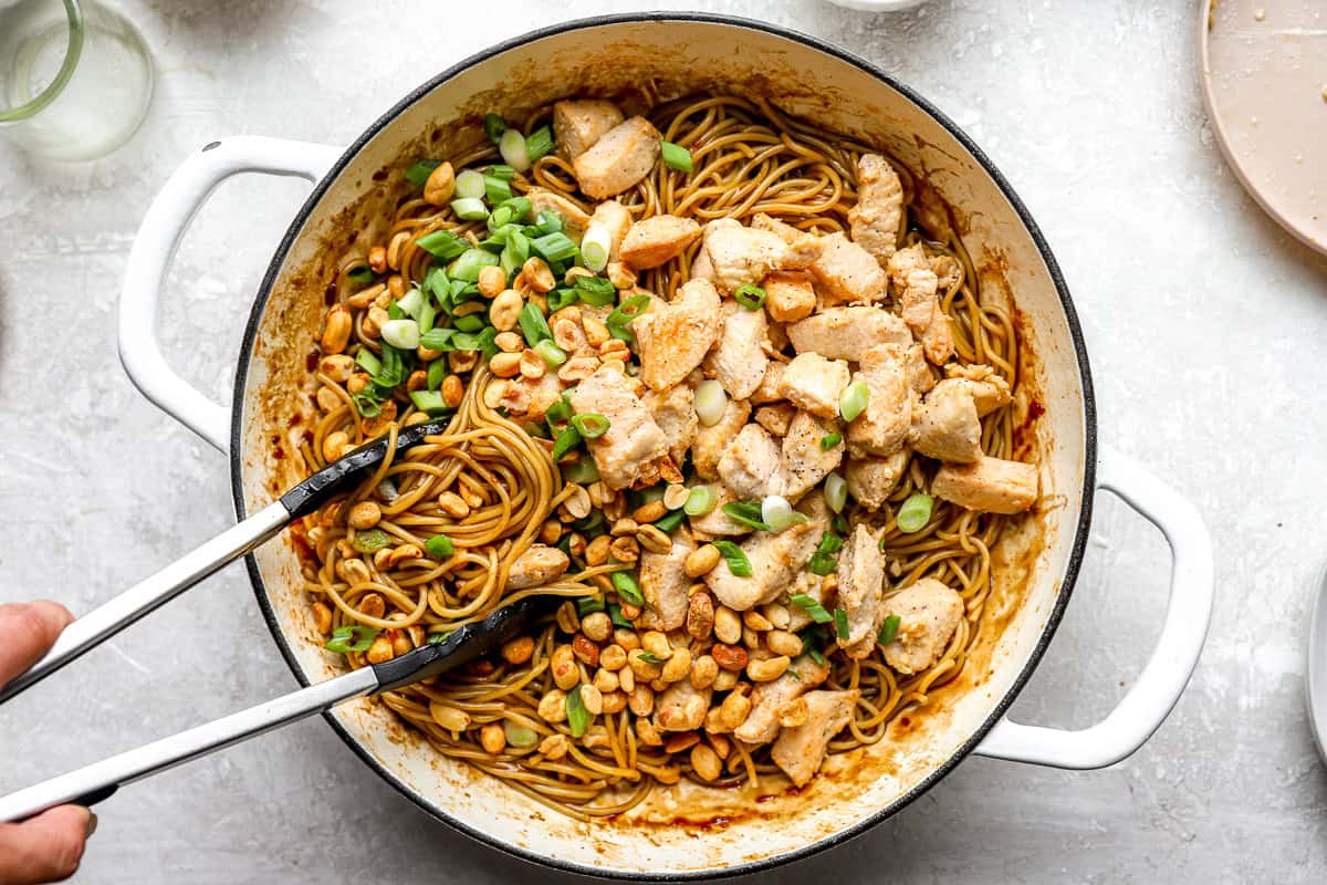 Kung pao spaghetti being tossed together with chicken, peanuts, and green onions in a skillet with tongs