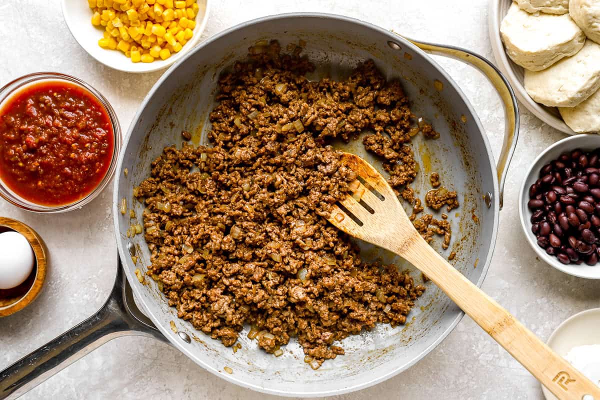 Ground beef and onions browning in a skillet, with bowls of corn, salsa, egg, and biscuit dough nearby.