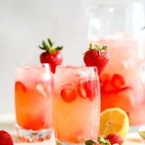 Two glasses of copycat Red Robin strawberry lemonade with sliced strawberries and ice, with a whole strawberry garnish and a pitcher behind them