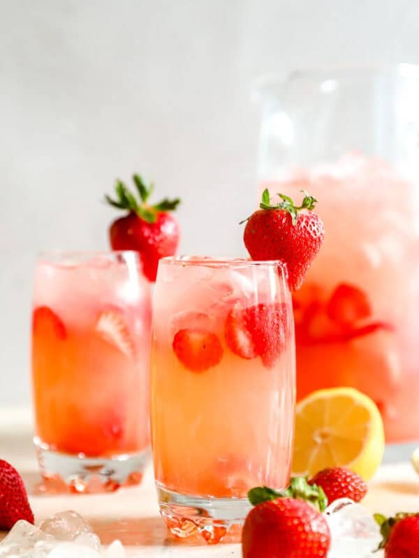 Two glasses of copycat Red Robin strawberry lemonade with sliced strawberries and ice, with a whole strawberry garnish and a pitcher behind them