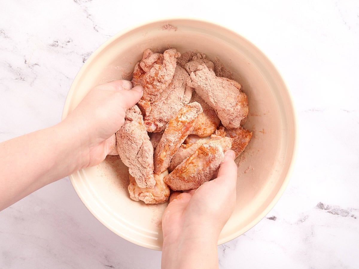 Hands tossing raw chicken wings in the seasoned flour mixture until fully coated.