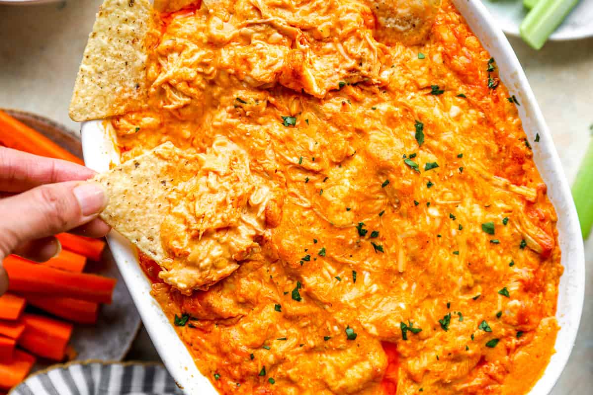 A hand dipping a tortilla chip into warm, creamy buffalo chicken dip.