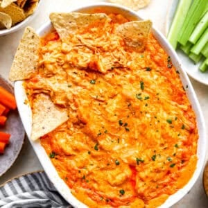 Baked buffalo chicken dip in a white oval baking dish, topped with tortilla chips and fresh herbs, with more chips, celery, and carrots alongside.