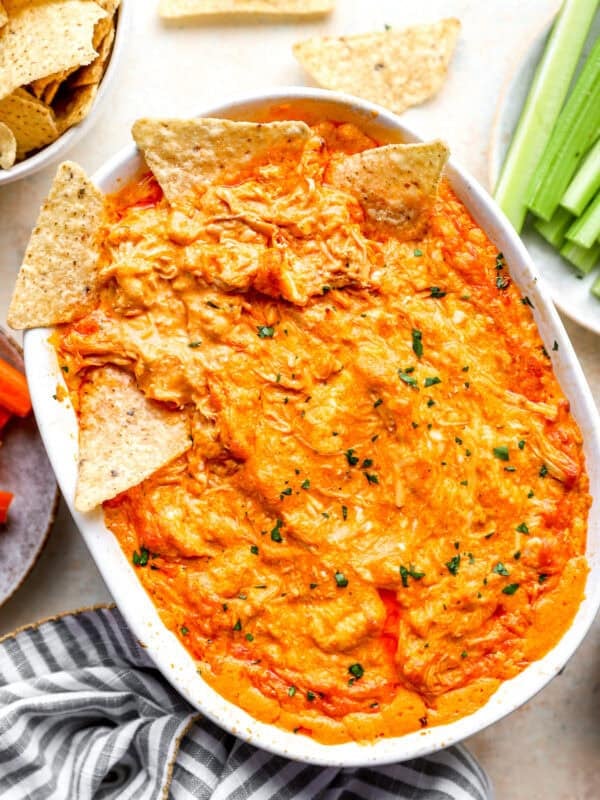 Baked buffalo chicken dip in a white oval baking dish, topped with tortilla chips and fresh herbs, with more chips, celery, and carrots alongside.