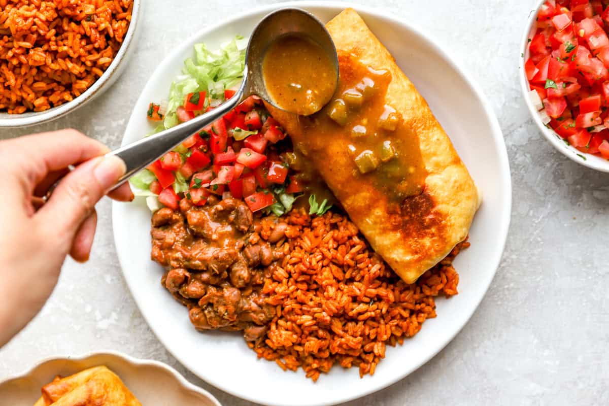 A spoon pouring warm green chile gravy over a fried chimichanga plated with Mexican rice, pinto beans, pico de gallo, and shredded lettuce.
