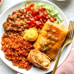 Overhead shot of a Chi-Chi's chimichanga cut in half on a white plate, showing shredded chicken and melted cheese inside, topped with green chile gravy and served with Mexican rice, pinto beans, cornbread, pico de gallo, and shredded lettuce.