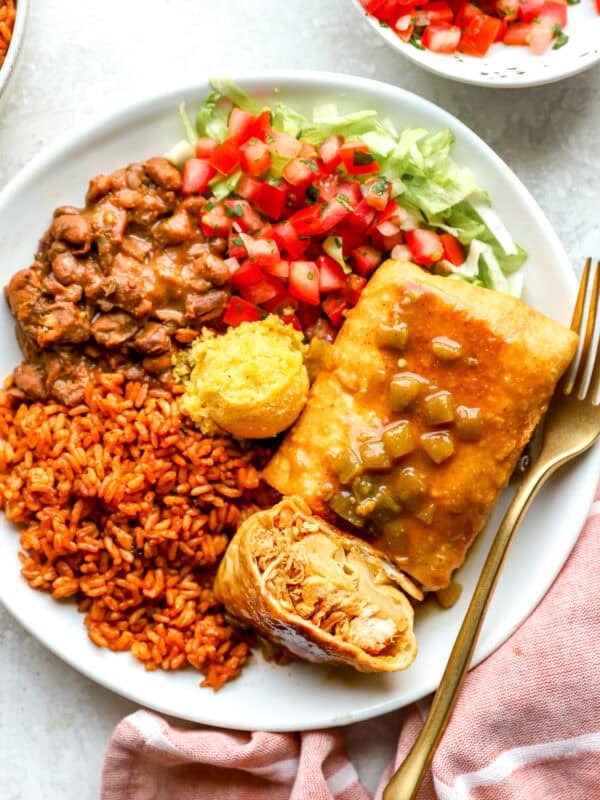 Overhead shot of a Chi-Chi's chimichanga cut in half on a white plate, showing shredded chicken and melted cheese inside, topped with green chile gravy and served with Mexican rice, pinto beans, cornbread, pico de gallo, and shredded lettuce.
