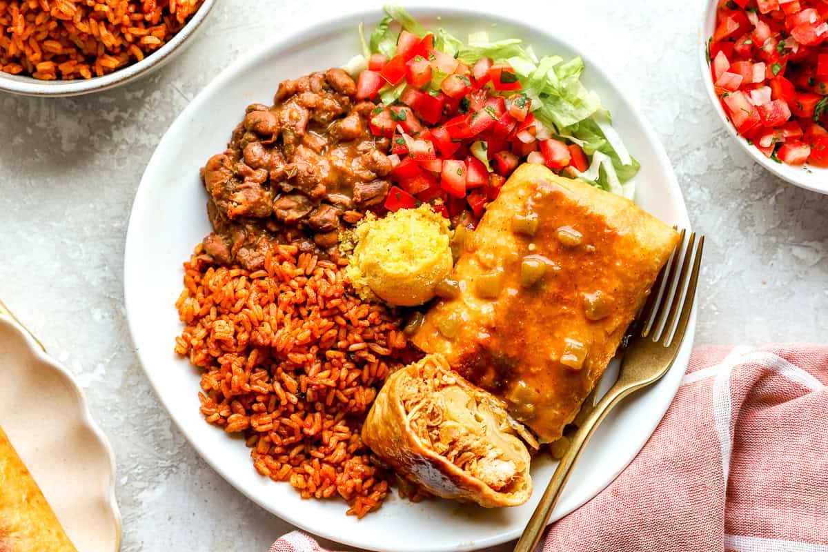 Close-up of a plated chimichanga with a forkful of shredded chicken pulled from the center, served alongside pinto beans, Mexican rice, cornbread, and pico de gallo.