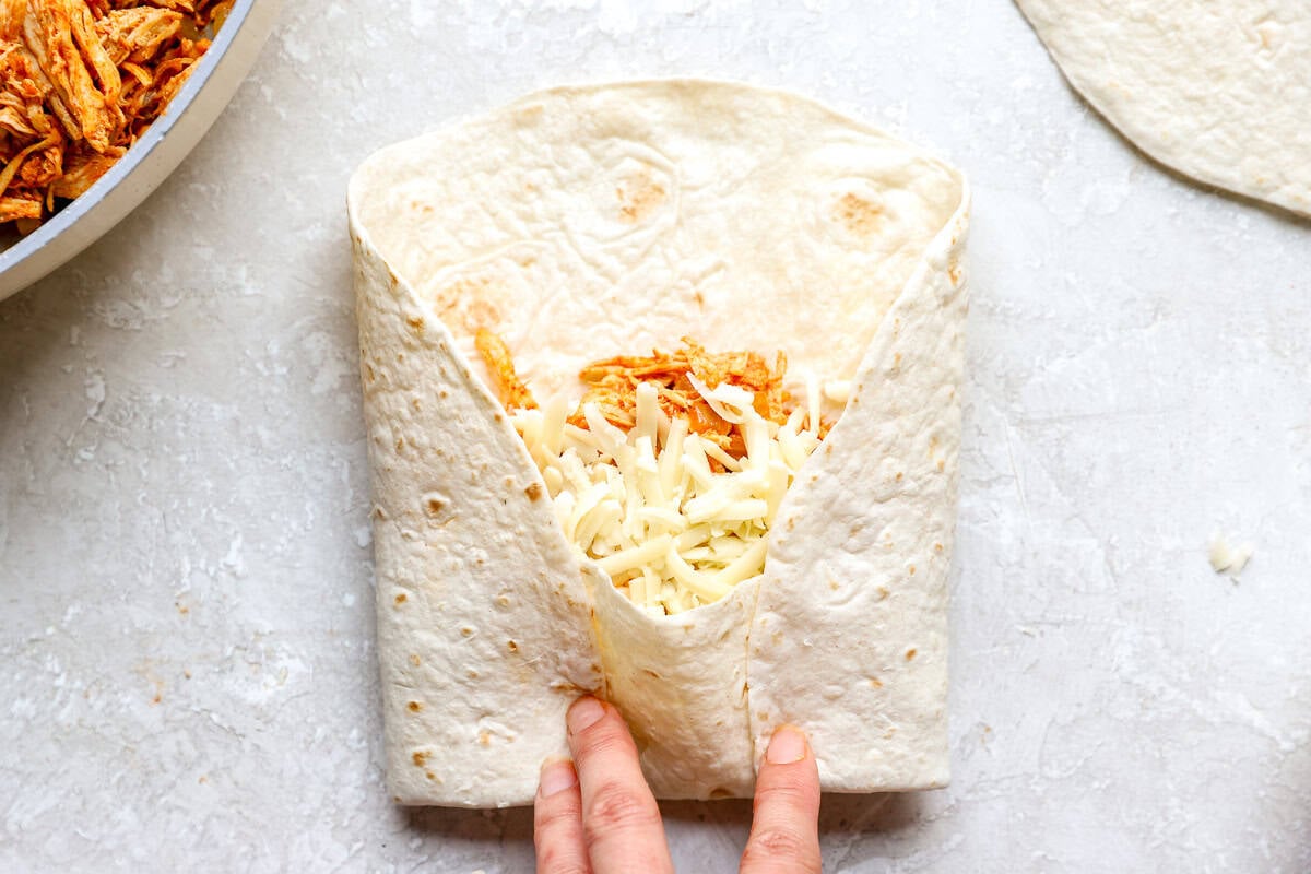 Hands folding the sides of a tortilla in over the chicken and cheese filling.