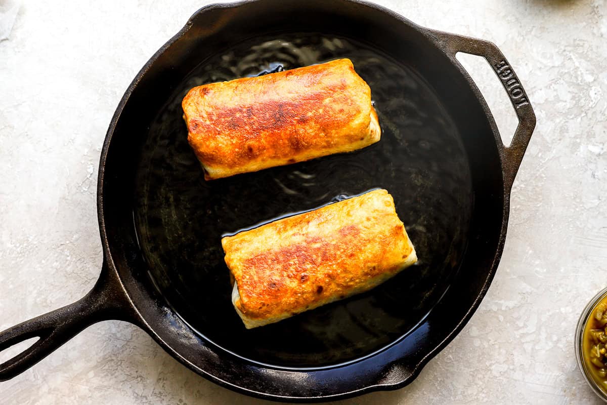 Two chimichangas frying in a cast iron skillet until golden brown and crispy.