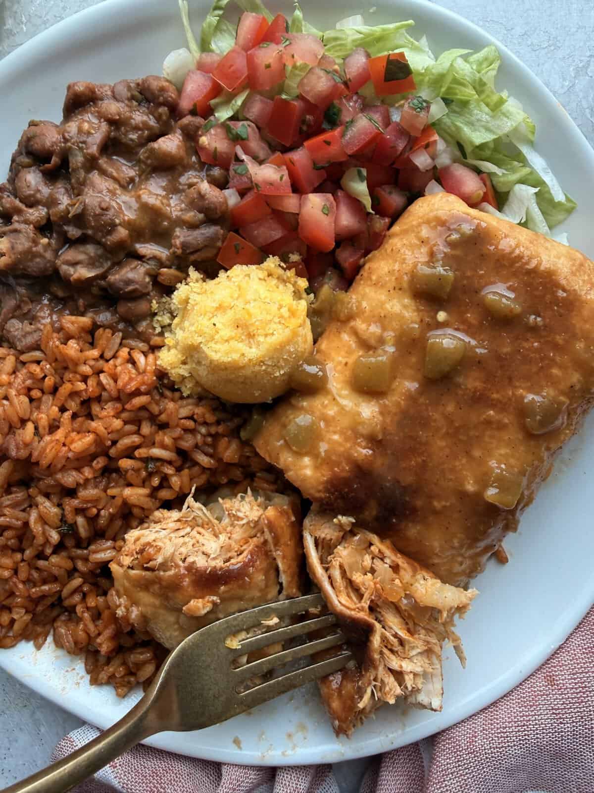 Close-up of a plated chimichanga with a forkful of shredded chicken pulled from the center, served alongside pinto beans, Mexican rice, cornbread, and pico de gallo.