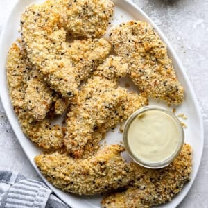 Everything bagel chicken strips piled on a white platter with a jar of creamy dipping sauce.