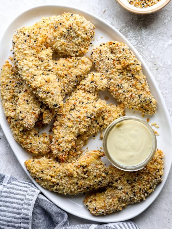 Everything bagel chicken strips piled on a white platter with a jar of creamy dipping sauce.