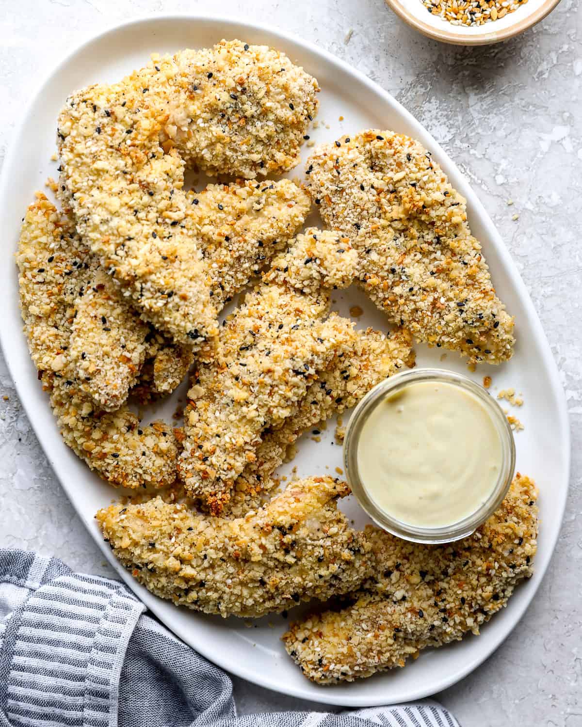 Everything bagel chicken strips piled on a white platter with a jar of creamy dipping sauce.