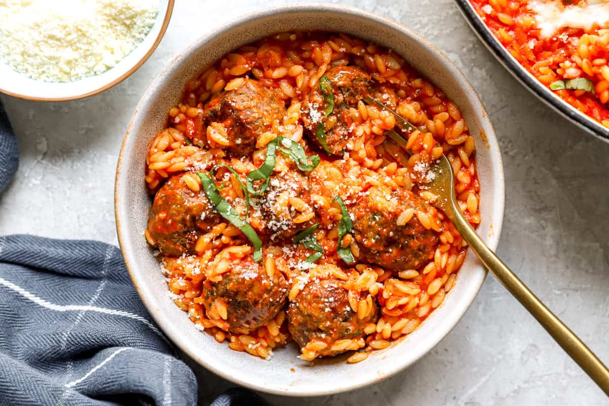 Close-up of meatball parmesan orzo in a bowl, garnished with basil and parmesan, fork twirling a bite.